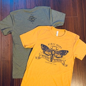 May include: Two t-shirts, one in a dark green color and one in a bright yellow color. The yellow t-shirt has a graphic of a moth with a skull on its head and the text "Piney Woods Mystic Menagerie Nacogdoches Texas".