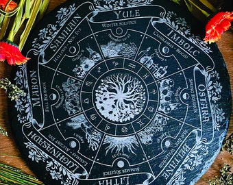 May include: A round, black slate board with a white engraved design featuring a tree of life at the center, surrounded by zodiac symbols and seasonal markers like Yule and Beltane. Floral accents and text complete the design.