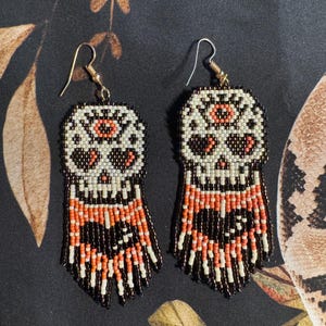 May include: A pair of beaded dangle earrings featuring a skull design with orange and white accents. The earrings have a black fringe.