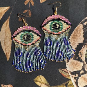 May include: A pair of beaded eye earrings with blue, green, and purple fringe. The earrings are made with small beads and have a gold hook.