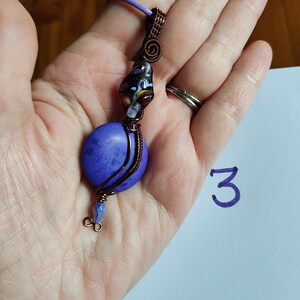 May include: A handcrafted pendant necklace featuring a large, round, purple stone. The stone is wire-wrapped with copper wire, and accented with a colorful glass bead. The necklace has a purple cord.