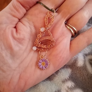 May include: A copper wire wrapped pendant with two white stones and a purple flower charm. The pendant is hanging from a green cord.