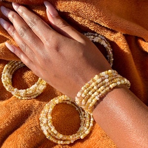 Puede incluir: Un conjunto de pulseras de cuentas en tonos amarillos y blancos. Las pulseras están hechas de pequeñas cuentas redondas y están apiladas unas sobre otras.