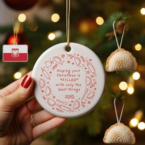 Puede incluir: Adorno navideño de cerámica blanca con un diseño de pierogi rojo y el texto "Hoping your Christmas is 'FILLED' with only the best things 2025." Otros adornos incluyen banderas polacas y decoraciones en forma de pierogi.