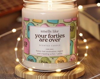 Op de afbeelding: Een brandende geurkaars in een glazen pot met de tekst "smells like your forties are over". De kaars heeft een kleurrijk label met grafsteenafbeeldingen en staat op een houten onderzetter. De geurnoten omvatten verlies van motivatie, spijt en pijn.