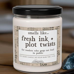May include: A white soy blend candle in a glass jar with a black lid. The label reads "smells like... fresh ink + plot twists" and includes text about the candle's burn time and scent. A feather and a blue stone are next to the candle.