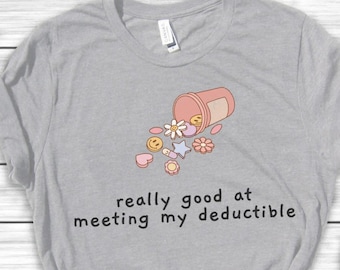 May include: A gray t-shirt with a pink pill bottle spilling out colorful pills with smiley faces, stars, and hearts. The text on the shirt reads "really good at meeting my deductible".