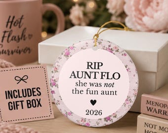 May include: A round ceramic ornament featuring a light pink center with the black text RIP AUNT FLO, she was not the fun aunt, a small black heart, and the year 2026. The border has a floral pattern with pink roses and green leaves.