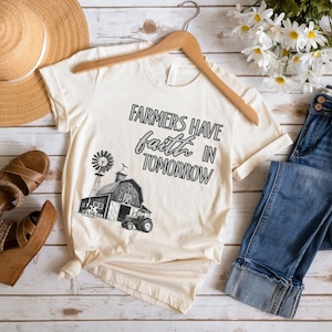 May include: A beige t-shirt with a black graphic of a farmhouse, windmill, and tractor. The text "Farmers have faith in tomorrow" is printed on the shirt.