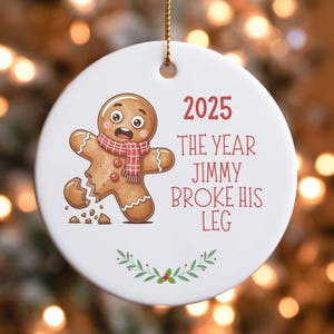 Op de afbeelding: Een witte ronde kerstbal met een afbeelding van een peperkoeken mannetje met een gebroken been. De tekst luidt "2025 The Year Jimmy Broke His Leg". Het peperkoeken mannetje draagt een rood-witte sjaal.