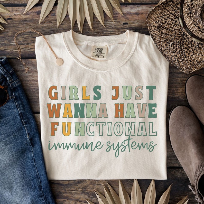 May include: A beige t-shirt with the text "Girls Just Wanna Have Functional Immune Systems" in colorful letters. The shirt is laid out on a wooden surface with a pair of brown shoes and a straw hat.