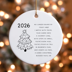 May include: A round white ceramic ornament with a gold hanging string. The front features the year 2026, a black line-art Christmas tree, and the text: NO LONGER BOUND BY FOGGY HAZE, I WATCH WITH PRIDE, YOUR SOBER DAYS, A JOURNEY LONG, WITH STEPS SO SMALL, YOU'VE RISEN HIGH, YOU'VE STOOD SO TALL, IN MOMENTS PURE, AND HEARTS SO FREE, A SOBER CHRISTMAS, BLESSED YOU'LL BE.