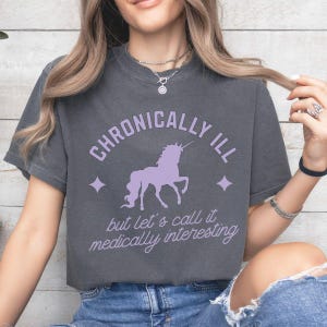 Puede incluir: Una camiseta gris con un gráfico de unicornio morado y el texto "Chronically Ill but let's call it medically interesting".