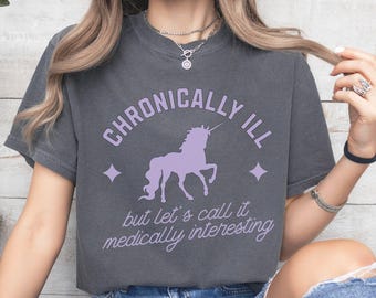 Może przedstawiać: Szary T-shirt z grafiką fioletowego jednorożca i tekstem "Chronically Ill but let's call it medically interesting".