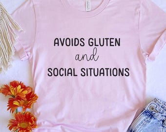 Op de afbeelding: Roze T-shirt met de tekst "Vermijdt Gluten en sociale situaties" in zwart gedrukt.