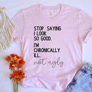 May include: Pink t-shirt with black text that reads "STOP SAYING I LOOK SO GOOD. I'M CHRONICALLY ILL. not ugly."