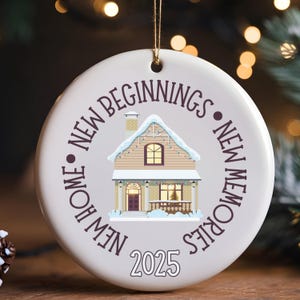 Könnte beinhalten: Runder, weißer Keramik-Weihnachtsschmuck mit einer Hausillustration. Das beige Haus hat ein schneebedecktes Dach, umgeben vom Text "New Home, New Beginnings, New Memories" und dem Jahr "2025". Ein goldener Faden ist befestigt.