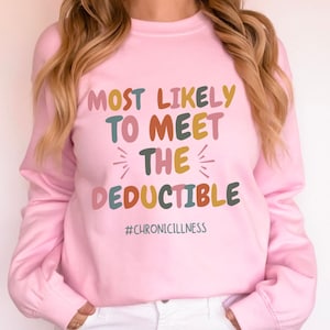 May include: A light pink sweatshirt with the text "MOST LIKELY TO MEET THE DEDUCTIBLE" in colorful letters. The hashtag "#CHRONICILLNESS" is below the text. The sweatshirt has long sleeves.