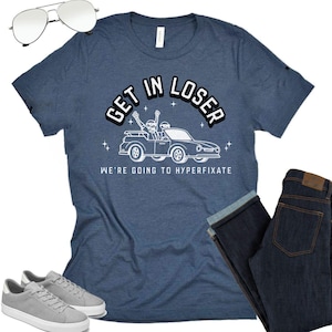 May include: A navy blue t-shirt with the text "GET IN LOSER" above a car graphic, and "WE'RE GOING TO HYPERFIXATE" below. The image also shows aviator sunglasses, dark wash jeans, and grey trainers.