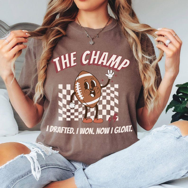 Peut inclure: Un t-shirt marron avec un personnage de football de dessin anim&eacute; et le texte "The Champ I drafted. I won. Now I gloat."