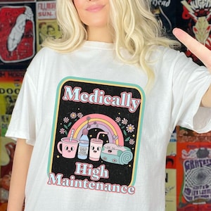 Puede incluir: Camiseta blanca con un gráfico colorido que dice "Medically High Maintenance". El diseño incluye un arcoíris, flores e ilustraciones de una taza, una botella de agua y una toalla enrollada. Una prenda casual para el día a día.