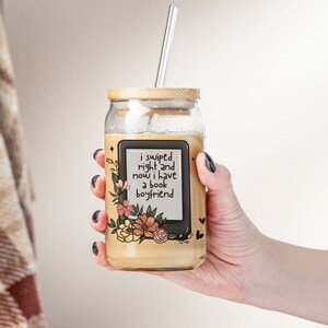 Peut inclure: Un bocal en verre avec un couvercle en bambou et une paille. Le bocal est décoré d'un motif floral et d'une image noir et blanc d'une liseuse avec le texte "i swiped right and now i have a book boyfriend".