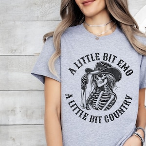 May include: Gray t-shirt with a black and white graphic of a skeleton wearing a cowboy hat. The text on the shirt reads "A little bit emo a little bit country".