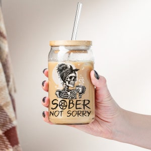 May include: A glass mason jar with a bamboo lid and a straw filled with a brown liquid. The jar has a black and white graphic of a skeleton with a bandana holding a cup of coffee. The text on the jar reads "SOBER NOT SORRY".