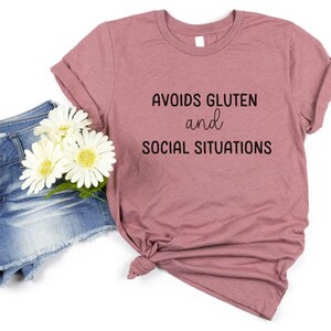 May include: A mauve t-shirt with the text "AVOIDS GLUTEN and SOCIAL SITUATIONS" printed in black.