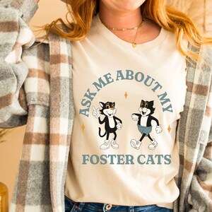 Könnte beinhalten: Ein weißes T-Shirt mit einem blauen und weißen Grafikdesign mit zwei Comic-Katzen und dem Text "Ask Me About My Foster Cats".