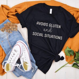 May include: Black t-shirt with white text that reads "Avoids Gluten and Social Situations". The shirt is laid out on a white wooden surface with a pair of white trainers, blue jeans, and a brown knitted scarf.