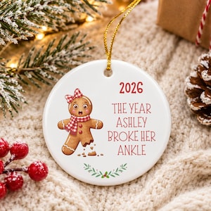 May include: A round white ceramic ornament featuring a gingerbread person with a red and white checkered bow and scarf, a broken leg, and the text 2026 THE YEAR ASHLEY BROKE HER ANKLE, finished with a gold hanging string.