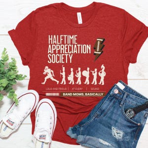 May include: Red t-shirt with the text "HALFTIME APPRECIATION SOCIETY" and a football graphic. The text "LOUD AND PROUD | AT EVERY | SOUND" is below the graphic. The text "BAND MOMS, BASICALLY" is at the bottom of the shirt.