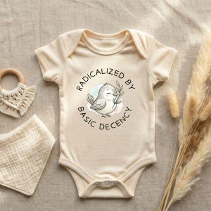 May include: Cream-colored baby bodysuit with the text "RADICALIZED BY BASIC DECENCY" surrounding a cartoon bird holding a sprig. A wooden teething ring and a muslin bib are also visible.