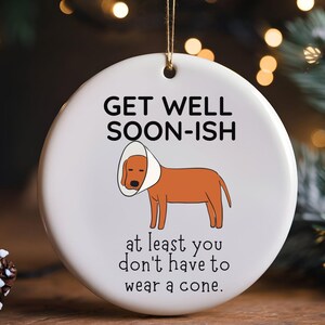 Könnte beinhalten: Runder weißer Keramikschmuck mit goldenem Faden, der einen braunen Hund mit Halskrause zeigt. Der Text lautet "GET WELL SOON-ISH" und "at least you don't have to wear a cone."