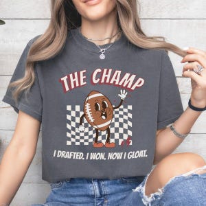 Peut inclure: Un t-shirt gris fonc&eacute; avec un personnage de football de dessin anim&eacute; portant des chaussures rouges et le texte "The Champ I Drafted. I Won. Now I Gloat."