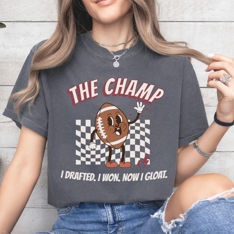 Peut inclure: Un t-shirt gris fonc&eacute; avec un personnage de football de dessin anim&eacute; portant des chaussures rouges et le texte "The Champ I Drafted. I Won. Now I Gloat."