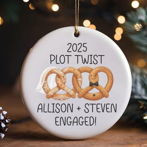 Può includere: Ornamento natalizio in ceramica bianca con cordino dorato. L'ornamento presenta il testo "2025 Plot Twist" e "Allison + Steven Engaged!". Sono raffigurati due personaggi pretzel, uno con velo e l'altro con papillon.