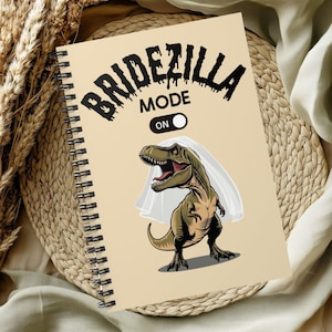 Puede incluir: Una libreta espiral beige con un dinosaurio con velo de novia y el texto "Bridezilla Mode On".
