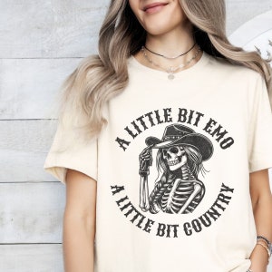 May include: A white t-shirt with a black graphic of a skeleton wearing a cowboy hat. The graphic says "A little bit emo, a little bit country".