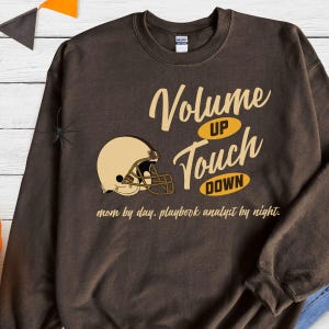 Op de afbeelding: Een bruine sweatshirt met een footballhelm-afbeelding en de woorden "Volume Up Touch Down". De tekst "mom by day. playbook analyst by night" is onder de afbeelding gedrukt. De sweatshirt is effen van kleur.