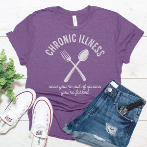 May include: Purple t-shirt with the text "CHRONIC ILLNESS" and a white fork and spoon crossed over each other. Below the text is the phrase "once you're out of spoons you're forked".