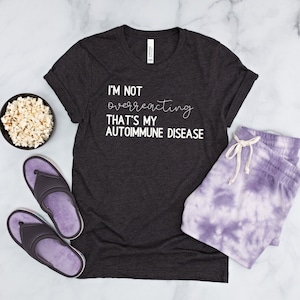 May include: A dark grey t-shirt with the text "I'm not overreacting that's my autoimmune disease" printed in white. The shirt is laid flat on a white marble surface with a bowl of popcorn, a pair of purple slippers, and a pair of purple tie-dye shorts.