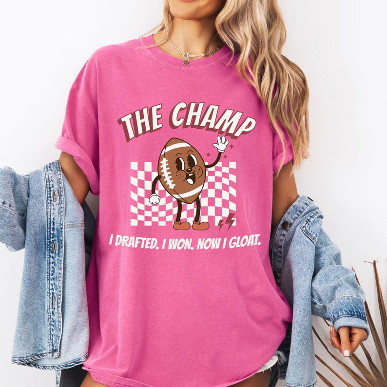 Peut inclure: T-shirt rose avec un dessin de football de bande dessin&eacute;e et le texte "The Champ I Drafted. I Won. Now I Gloat."