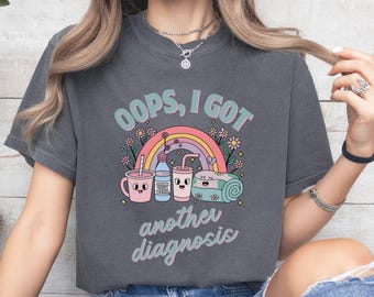 May include: Dark gray t-shirt with the text "OOPS, I GOT another diagnosis" in a whimsical font. The graphic includes a rainbow, flowers, and cartoon illustrations of a drink cup, water bottle, and rolled towel.
