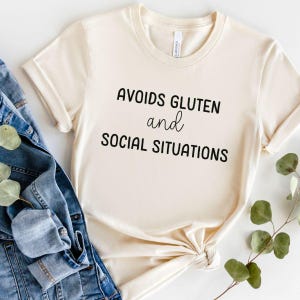 May include: A beige t-shirt with black text that reads "Avoids Gluten and Social Situations".