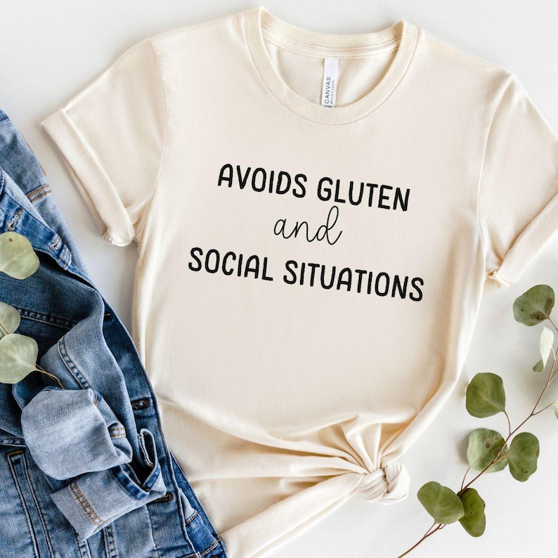 May include: A beige t-shirt with black text that reads "Avoids Gluten and Social Situations".