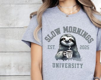 Pode incluir: Camiseta cinza com um gráfico de um bicho-preguiça segurando uma caneca de café que diz "Awake but at what cost?" O gráfico também inclui o texto "Slow Mornings University Est. 2025".