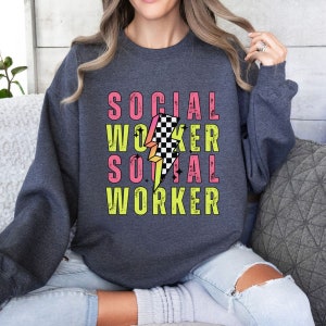 Könnte beinhalten: Ein dunkelgrauer Pullover mit dem Text "SOCIAL WORKER", der zweimal in Pink und Gelb wiederholt wird, mit einem pinkfarbenen und gelben Blitzgrafik mit einem schwarz-weiß karierten Muster in der Mitte.