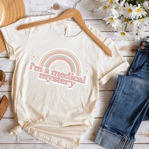 May include: A beige t-shirt with a rainbow and the text "i'm a medical mystery" in pink.
